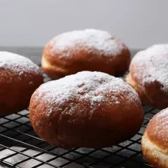Pięć pysznych pączków z piekarnika, oprószonych cukrem pudrem, leży na czarnej kratce.