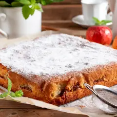 Puszysty biszkopt z jabłkami, posypany cukrem pudrem, obok jabłka i laski cynamonu. Idealny na deser.
