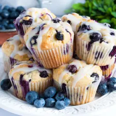 Stos świeżo upieczonych muffinek z borówkami, polanych lukrem, z dodatkiem świeżych owoców na talerzu.