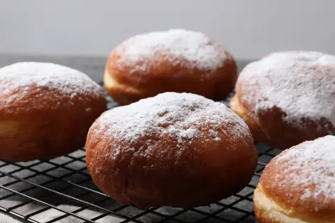 Pięć pysznych pączków z piekarnika, oprószonych cukrem pudrem, leży na czarnej kratce.
