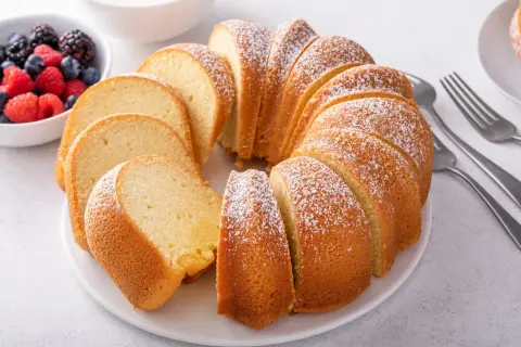 Wilgotna babka piaskowa, oprószona cukrem pudrem, prezentuje się apetycznie obok miseczki z jagodami i malinami.