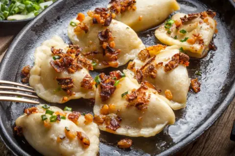 Pierogi z serem, przygotowane według przepisu babci, polane skwarkami i szczypiorkiem, na czarnej patelni.