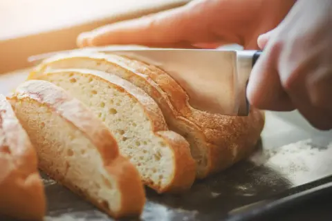 Dłoń kroi świeży chleb na kromki. Dowiedz się, jak odświeżyć chleb, aby był jak najsmaczniejszy.