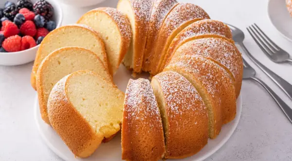 Wilgotna babka piaskowa, oprószona cukrem pudrem, prezentuje się apetycznie obok miseczki z jagodami i malinami.