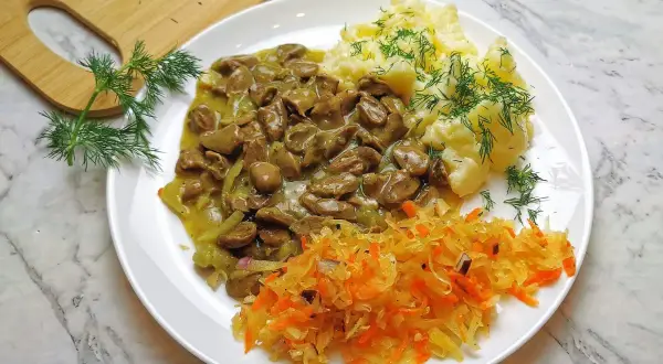 Pyszny gulasz z serc drobiowych z puree ziemniaczanym i surówką z kiszonej kapusty, ozdobiony świeżym koperkiem.
