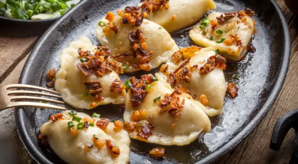 Pierogi z serem, przygotowane według przepisu babci, polane skwarkami i szczypiorkiem, na czarnej patelni.