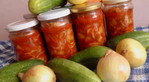 Słoiki z sałatką z ogórków z przecierem pomidorowym, obok świeże ogórki i cebule.