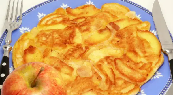Złocisty omlet z jabłkiem, podany na niebieskim talerzu z widelcem i nożem. Obok leży świeże jabłko.