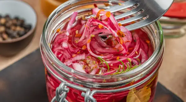 Słoik pełen aromatycznej, czerwonej cebuli marynowanej z ziołami. Widelec nabiera pyszne, różowe plasterki.