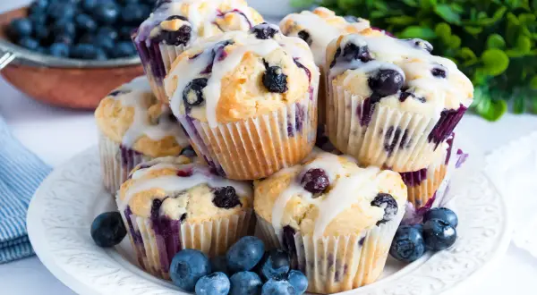 Stos świeżo upieczonych muffinek z borówkami, polanych lukrem, z dodatkiem świeżych owoców na talerzu.