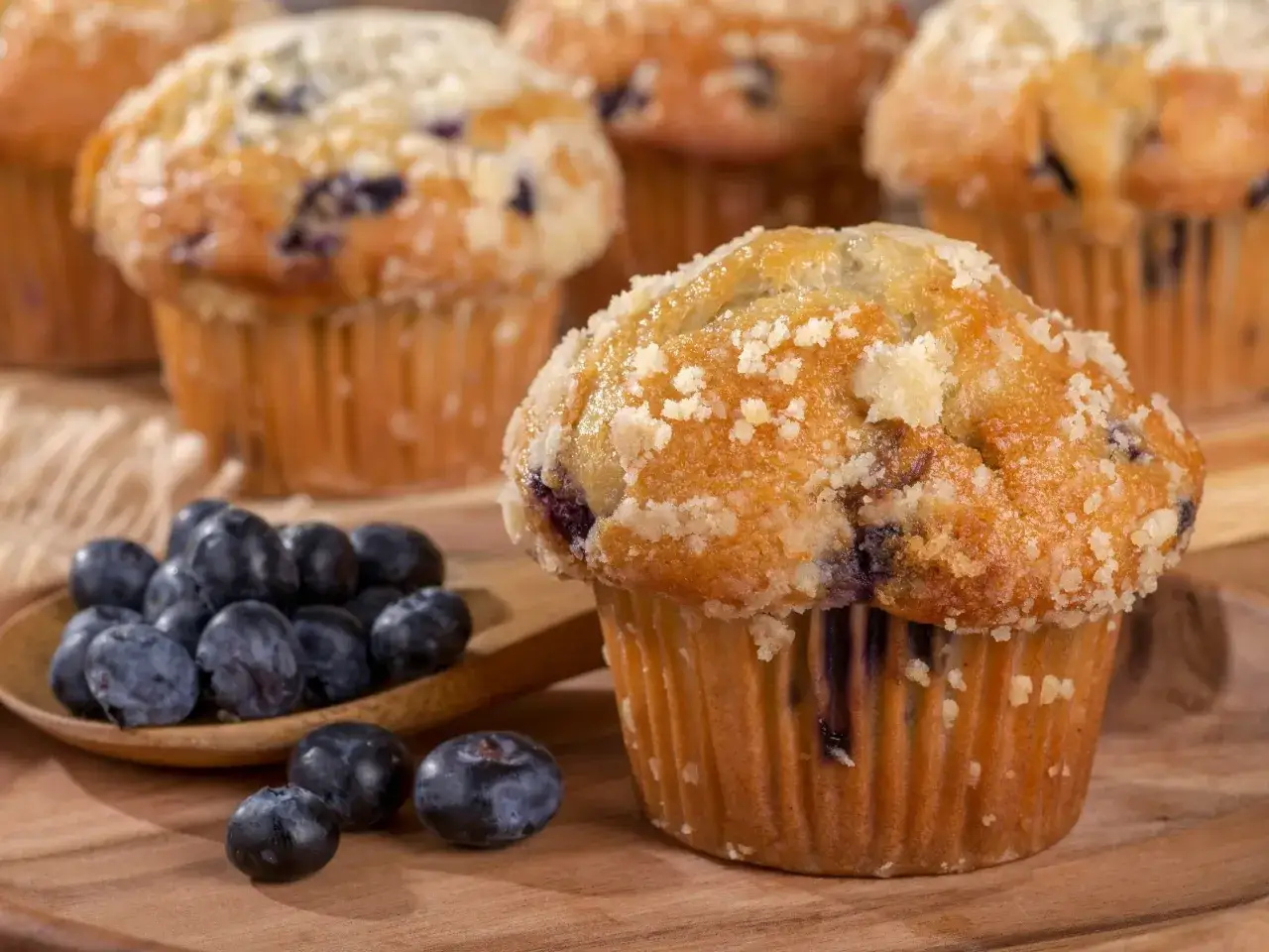 Puszyste muffinki z bor&oacute;wkami, posypane kruszonką, obok świeżych owoc&oacute;w na drewnianej desce.