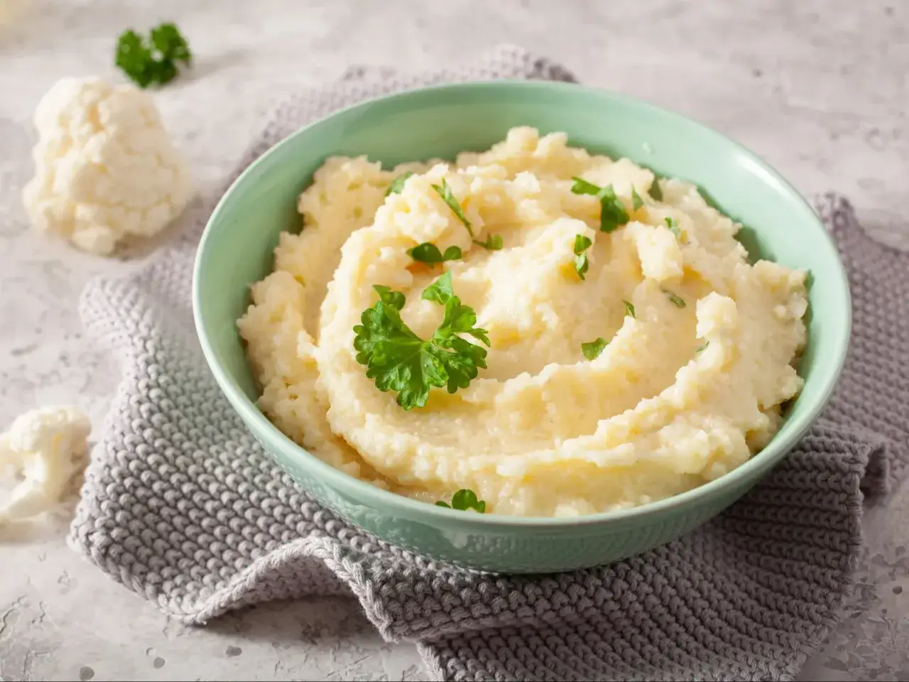 Aksamitne puree z kalafiora, udekorowane natką pietruszki, podane w miętowej misce na szarym, dzianinowym obrusie. Obok leżą świeże r&oacute;życzki kalafiora.