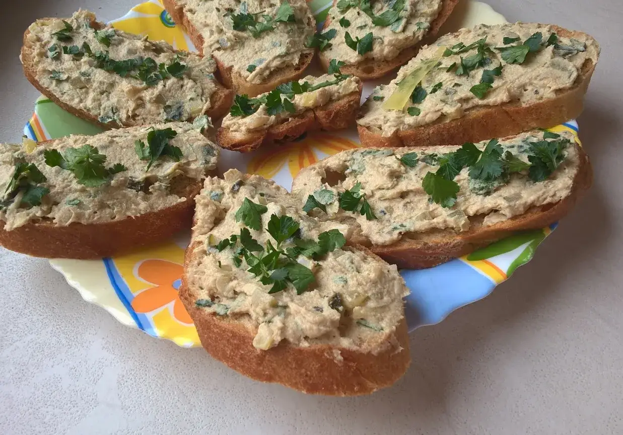 Kromki chleba z pastą z makreli, posypane natką pietruszki, ułożone na kolorowym talerzu.