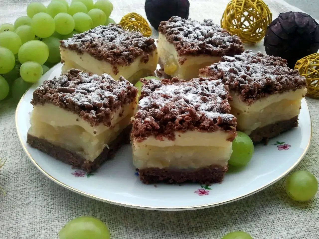 Szybkie ciasto z gruszkami i budyniem, posypane cukrem pudrem, prezentuje się apetycznie na talerzu, obok świeżych winogron.