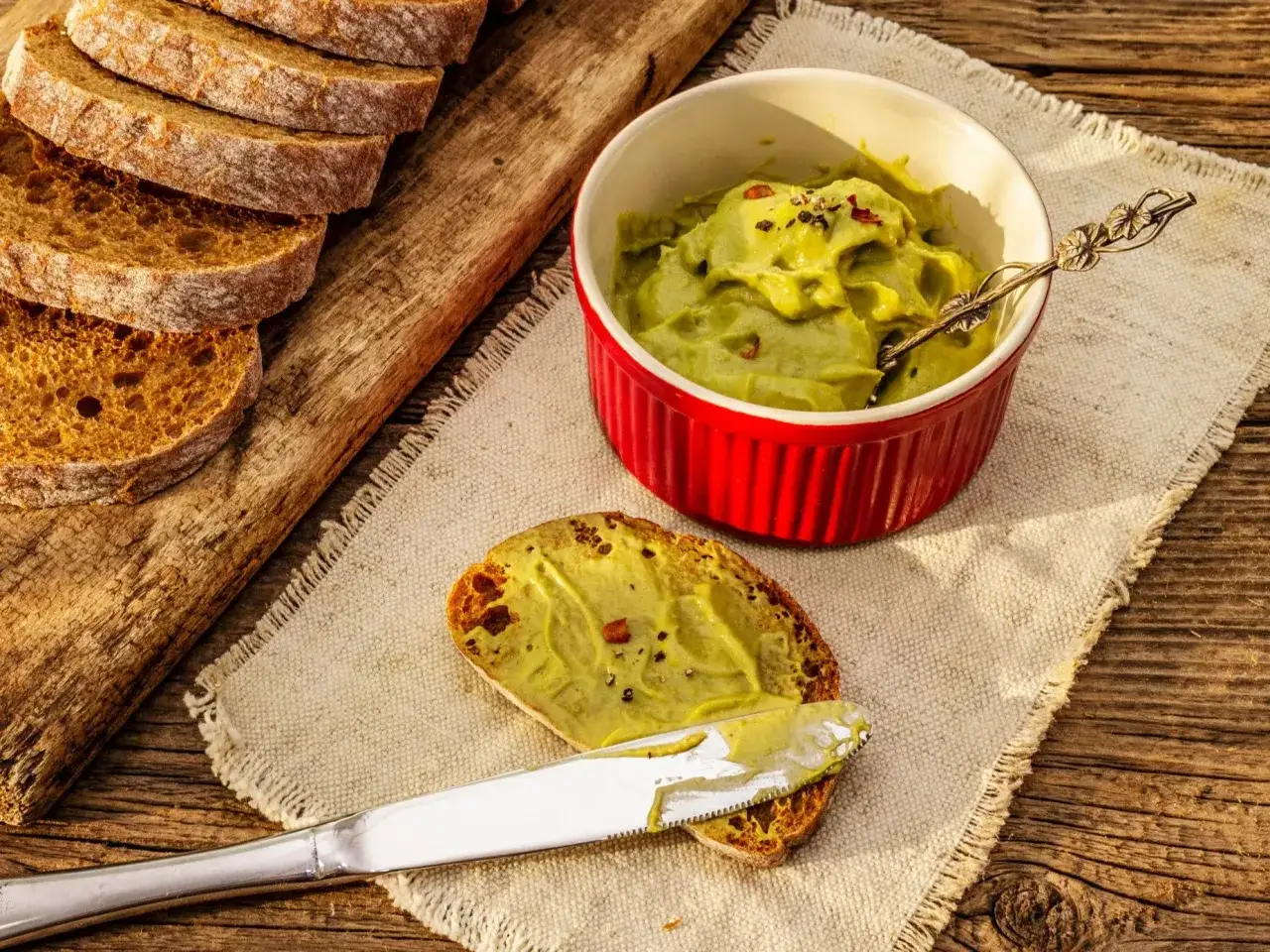 Pyszna pasta z awokado na kromce chleba, obok miseczka z pastą i kromki chleba na desce.