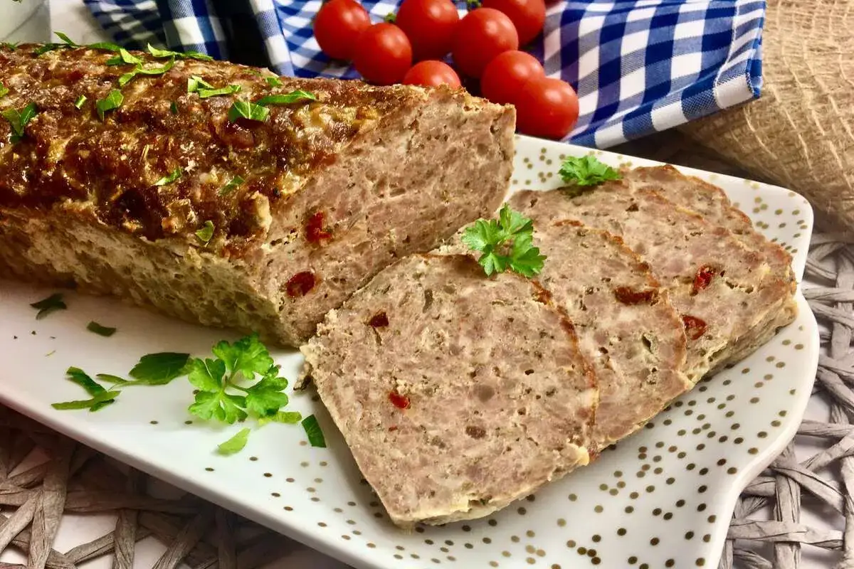Pyszna pieczeń z mięsa mielonego, pokrojona w plastry, ozdobiona natką pietruszki. Obok pomidorki i niebiesko-biała kratka.