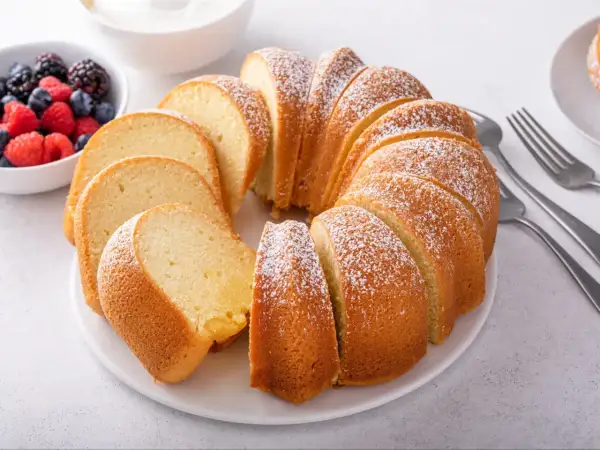 Wilgotna babka piaskowa, oprószona cukrem pudrem, prezentuje się apetycznie obok miseczki z jagodami i malinami.