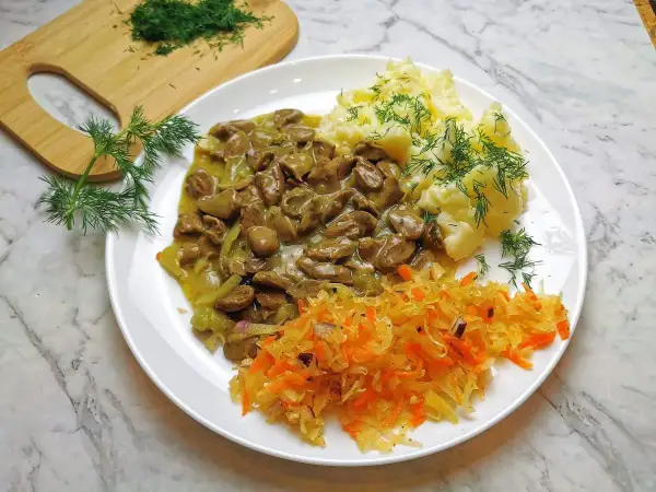 Pyszny gulasz z serc drobiowych z puree ziemniaczanym i surówką z kiszonej kapusty, ozdobiony świeżym koperkiem.